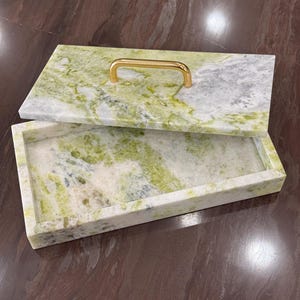 May include: A white marble tray with a green marble lid and a gold handle. The tray is rectangular and has a smooth surface. The lid has a swirling pattern of green and white.