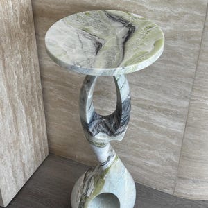 May include: A decorative side table with a round top and a unique, sculpted base. The table is made of marble with a green, white, and black color scheme. The base has an open, curved design. The table is approximately 20 inches tall.