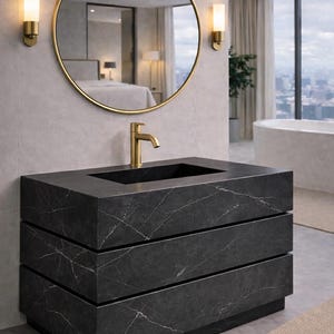 May include: A black marble vanity cabinet with two drawers and a rectangular sink basin. The vanity features a gold faucet, a large round mirror with a gold frame, and two gold wall sconces mounted on a light gray tiled wall above the unit.
