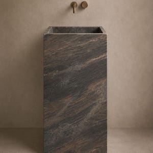 Puede incluir: Un lavabo de pedestal rectangular hecho de piedra de color marr&oacute;n oscuro y negro con vetas naturales prominentes. El lavabo est&aacute; integrado en la parte superior de la estructura, acompa&ntilde;ado por un grifo y mando de pared en bronce.
