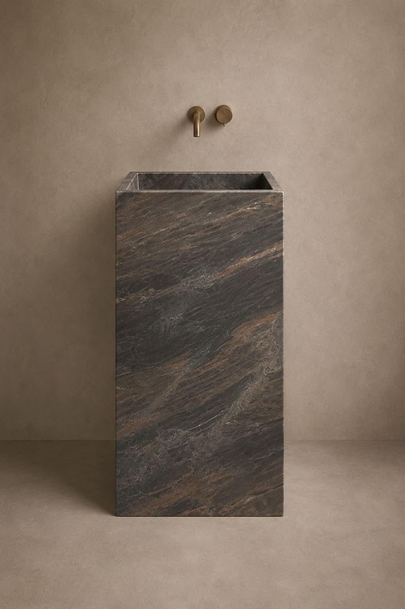 Puede incluir: Un lavabo de pedestal rectangular hecho de piedra de color marr&oacute;n oscuro y negro con vetas naturales prominentes. El lavabo est&aacute; integrado en la parte superior de la estructura, acompa&ntilde;ado por un grifo y mando de pared en bronce.