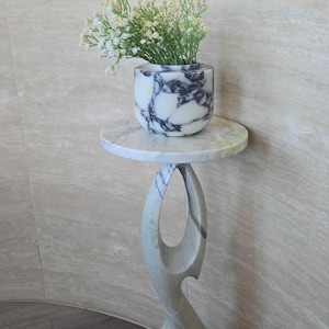 May include: A white and grey marble pedestal table with a circular top and a sculptural, hollowed-out base. A matching white and grey marble bowl sits on the table, containing a bouquet of small white flowers with green stems and foliage.