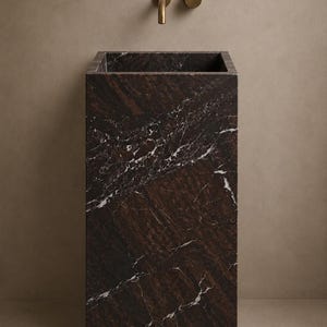 Puede incluir: Un lavabo de pedestal rectangular hecho de m&aacute;rmol marr&oacute;n oscuro con vetas blancas prominentes. El cuenco presenta una abertura cuadrada profunda. Montado en la pared sobre el lavabo hay un grifo de lat&oacute;n minimalista y un mando circular a juego.