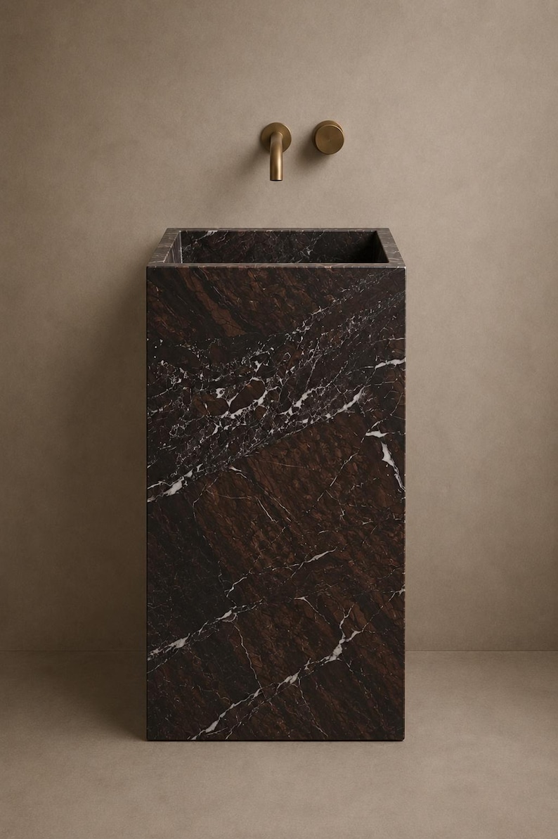 Puede incluir: Un lavabo de pedestal rectangular hecho de m&aacute;rmol marr&oacute;n oscuro con vetas blancas prominentes. El cuenco presenta una abertura cuadrada profunda. Montado en la pared sobre el lavabo hay un grifo de lat&oacute;n minimalista y un mando circular a juego.