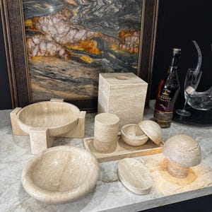 Pode incluir: Uma coleção de artigos de decoração para casa em pedra bege, incluindo tigelas, uma caixa de lenços, uma bandeja e um objeto em forma de cogumelo. Uma obra de arte abstrata emoldurada e uma garrafa de conhaque estão no fundo. Os itens estão dispostos em uma bancada de mármore.