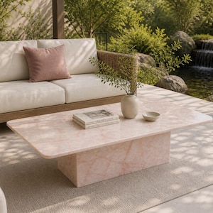 May include: A modern outdoor living space featuring a light pink rectangular coffee table with rounded corners. A beige sofa with a mauve pillow sits behind the table. A small vase, bowl, and book are on the table. A waterfall and greenery are in the background.