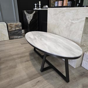 May include: Oval marble-topped coffee table with a black metal base. The tabletop features a light grey and white veined pattern. The table is a modern design, suitable for a living room or showroom.