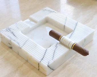 May include: A square white marble ashtray featuring grey veining and a ribbed texture on the top surface. A brown cigar with a gold and white paper band rests across one of the four indented notches designed for holding cigars or cigarettes.