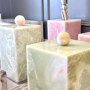 May include: Two large light green onyx cubes with polished surfaces, each topped with a small sphere. One sphere is light pink, the other is beige. A pink onyx cube stands behind them, topped with a tall, cylindrical, black and white striped stone object.