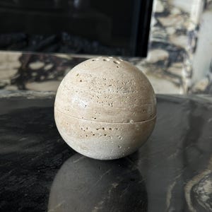 May include: A spherical, beige stone container with a textured surface and small holes on top. The container is split in half. The object is sitting on a dark, reflective surface.