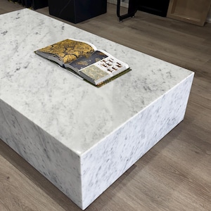 Könnte beinhalten: Ein rechteckiger Couchtisch aus weißem Marmor mit grauer Maserung. Ein offenes Buch mit goldenen und dunkelgrauen Seiten liegt auf dem Tisch. Der Tisch steht auf einem Holzboden.