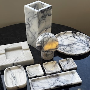 Peut inclure: Une collection d'articles de décoration intérieure en marbre blanc avec des veines grises. Les articles comprennent un grand vase carré, une sculpture en forme de champignon, une assiette ronde, un cendrier et plusieurs petits plateaux carrés et rectangulaires.
