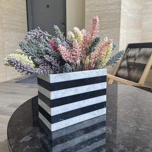 May include: A square marble vase with black and white horizontal stripes, filled with colorful faux flowers in shades of pink, yellow, green, and purple. The vase sits on a dark, round table.