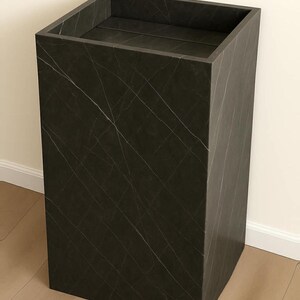 Puede incluir: Un pedestal o columna rectangular alta con un acabado gris carb&oacute;n oscuro que presenta vetas blancas finas y sutiles en toda la superficie. La estructura tiene una parte superior plana y abierta con bordes limpios y definidos.