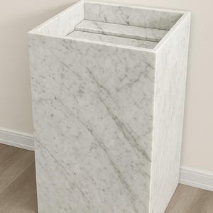 Puede incluir: Un lavabo de pedestal rectangular hecho de m&aacute;rmol blanco con vetas grises. La estructura presenta una secci&oacute;n superior hueca con un estante interno plano, mostrando una superficie de piedra lisa y pulida en todo el marco exterior.