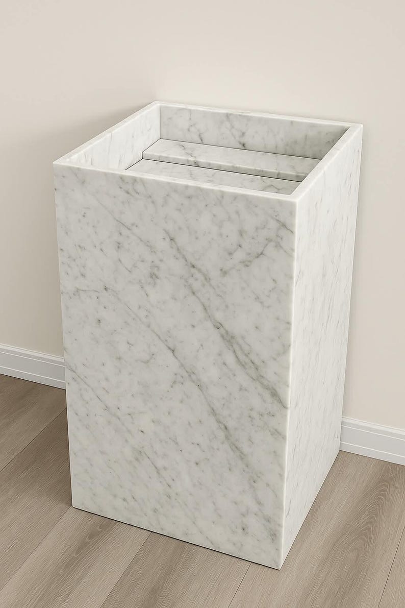 Puede incluir: Un lavabo de pedestal rectangular hecho de m&aacute;rmol blanco con vetas grises. La estructura presenta una secci&oacute;n superior hueca con un estante interno plano, mostrando una superficie de piedra lisa y pulida en todo el marco exterior.