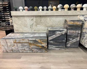 May include: A collection of stone furniture including a low rectangular coffee table, a square side table, and a tall rectangular pedestal, all featuring a dark grey, white, and gold veined marble pattern. A row of small mushroom-shaped stone decor sits atop a large marble counter.