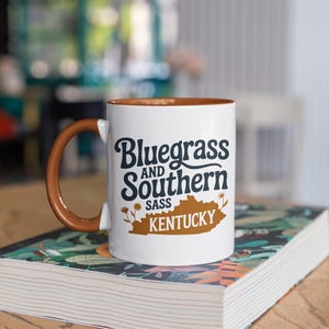 a coffee mug that says "bluegrass and southern sass Kentucky" with a kentucky and floral graphics. The design is part of A collection of Kentucky themed vector images perfect for cricut, silhouette, tshirt design, DTF Printing, POD, mugs, tote bags, sublimation and more!