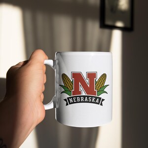 A white mug that says "nebraska" with an "N" graphic and two corn graphics