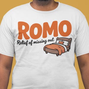 以下が含まれることがあります： オレンジ色のブロック文字で「ROMO」と書かれた白いTシャツ。「Relief of missing out」というフレーズと、オレンジ色の寝具の漫画のベッドが下にあります。