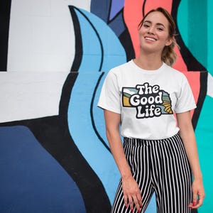 A girl standing against a colorful wall with a shirt that says "the good life"