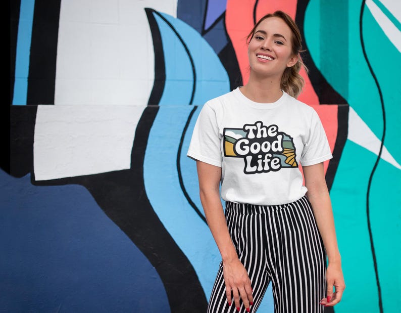 A girl standing against a colorful wall with a shirt that says "the good life"