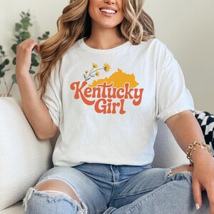 a woman wearing a white tshirt that says "kentucky girl" with a kentucky state silhouette and goldenrod graphics. The design is part of A collection of Kentucky themed vector images perfect for cricut, silhouette, tshirt design, DTF Printing, POD, mugs, tote bags, sublimation and more!