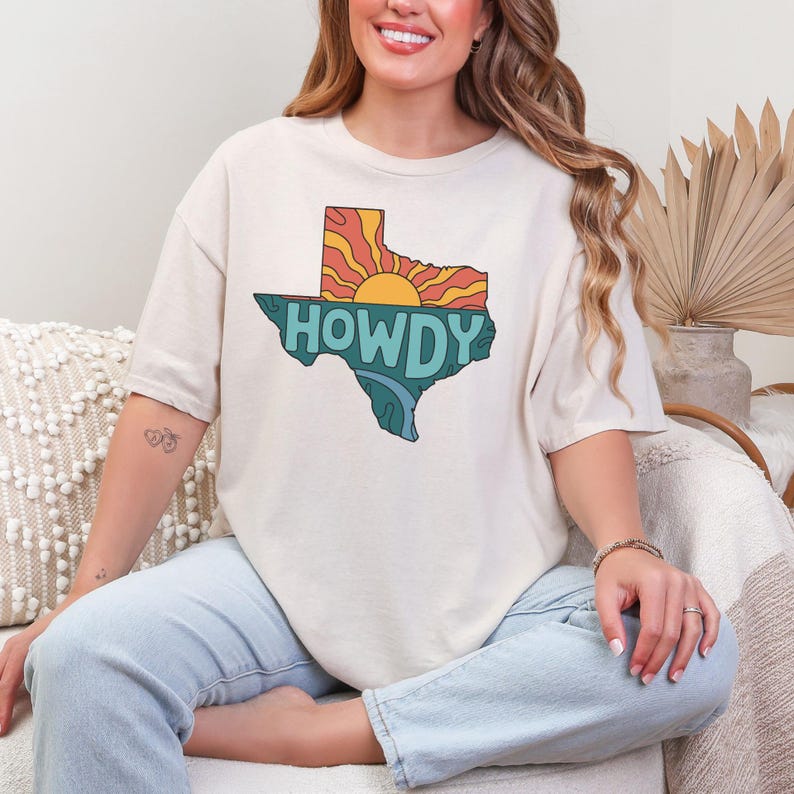 A woman sitting in front of an aesthetic beige backdrop wearing a white tshirt that says "Howdy" on a stylized texas silhouette