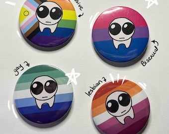 May include: Four round buttons with different pride flags and a cartoon alien face on each. The flags are labeled "Progressive", "Bisexual", "Gay", and "Lesbian".