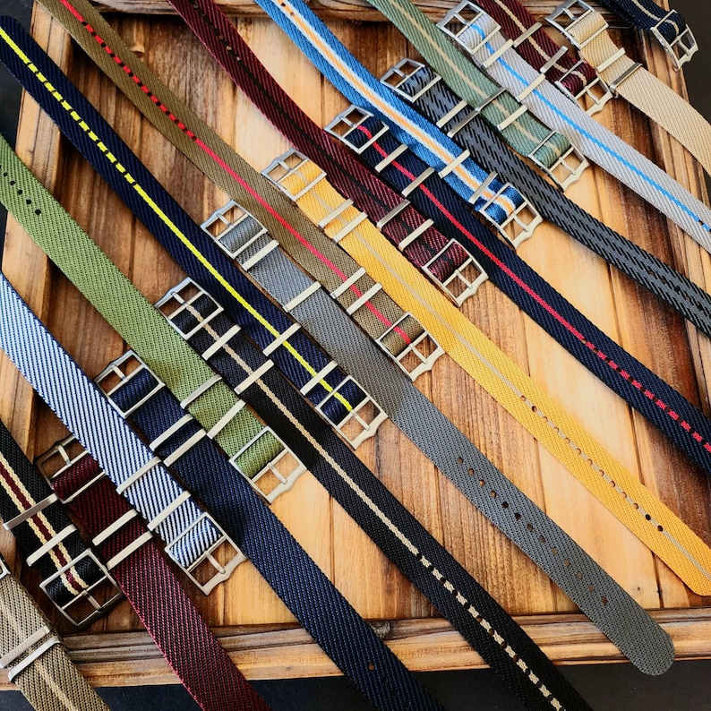 Pu&ograve; includere: Una collezione di cinturini colorati per orologi con vari disegni a righe e tinta unita. I cinturini sono realizzati in tessuto intrecciato e presentano fibbie color argento. I colori includono blu, verde, rosso, giallo e grigio. I cinturini sono disposti diagonalmente su una superficie di legno.