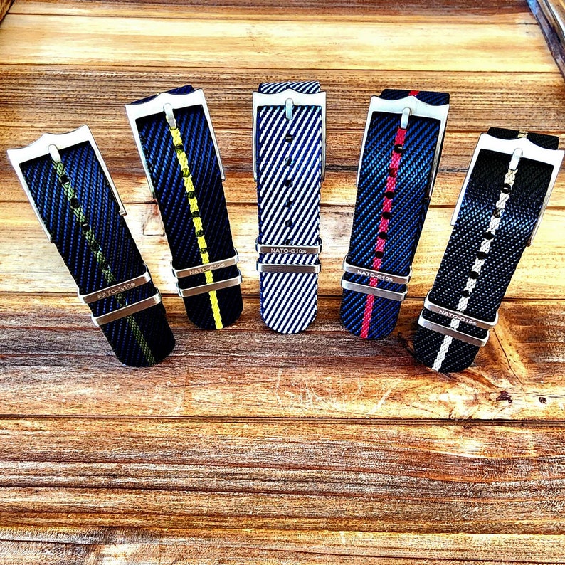 Pu&ograve; includere: Cinque cinturini per orologi in nylon con fibbie in acciaio inossidabile sono esposti su una superficie di legno. I cinturini presentano varie combinazioni di colori, tra cui blu con strisce gialle, rosse e bianche, e nero con dettagli bianchi. Ogni cinturino ha il testo "NATO-G10S".
