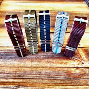 Pu&ograve; includere: Cinque cinturini per orologi in vari colori, tra cui rosso, verde, blu e grigio, sono esposti su una superficie di legno. Ogni cinturino presenta una fibbia argentata e un design intrecciato. I cinturini sono angolati, mostrando i loro motivi e le loro trame.