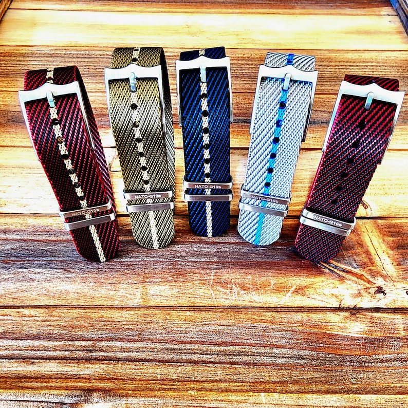 Pu&ograve; includere: Cinque cinturini per orologi in vari colori, tra cui rosso, verde, blu e grigio, sono esposti su una superficie di legno. Ogni cinturino presenta una fibbia argentata e un design intrecciato. I cinturini sono angolati, mostrando i loro motivi e le loro trame.