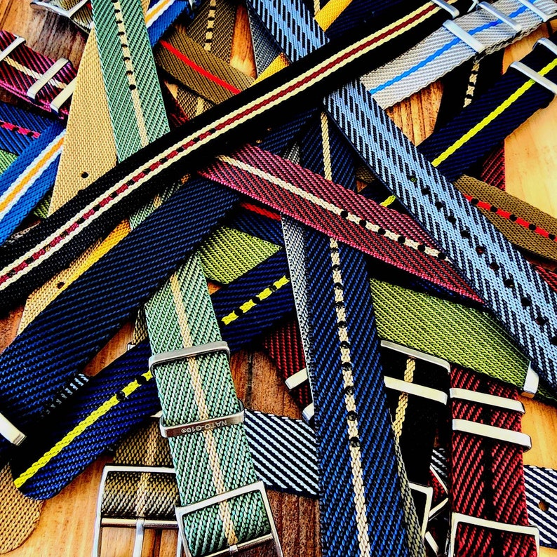 Pu&ograve; includere: Una collezione di cinturini colorati per orologi con vari disegni a righe e motivi. I cinturini sono in tonalit&agrave; di blu, verde, rosso e nero, con fibbie in metallo. I cinturini sono realizzati in tessuto intrecciato.