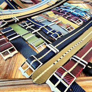 Pu&ograve; includere: Una collezione di cinturini per orologi di vari colori e motivi. I cinturini presentano una variet&agrave; di design intrecciati, tra cui strisce e colori uniti, con fibbie color argento. I cinturini sono disposti su una superficie di legno.