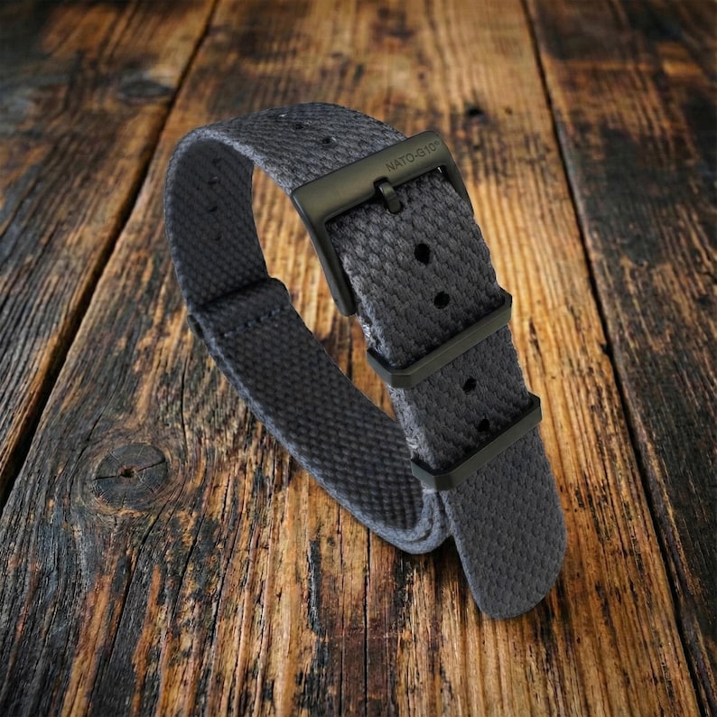 Puede incluir: Una correa de reloj gris estilo OTAN con una hebilla negra. La correa est&aacute; hecha de una tela texturizada y presenta el texto "NATO-G10" en la hebilla. La correa del reloj se muestra sobre una superficie de madera.