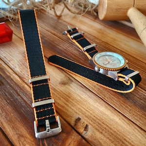 May include: Black watch strap with orange stitching and a silver buckle. The strap is made of nylon and is designed for a watch with a 20mm lug width.