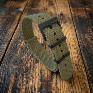 Puede incluir: Correa de reloj verde oliva con hebilla y presillas negras. La correa est&aacute; hecha de tela texturizada y se muestra sobre una superficie de madera. La correa del reloj tiene el texto "WATCHBAND" en la hebilla.