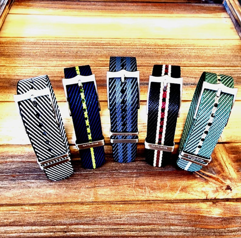 Pu&ograve; includere: Cinque cinturini per orologi in nylon con fibbie in acciaio inossidabile sono esposti su una superficie di legno. I cinturini presentano varie combinazioni di colori, tra cui strisce bianche e nere, blu con accenti gialli, strisce blu, nero con strisce rosse e bianche e verde con strisce bianche.