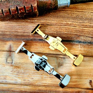 May include: Two watch clasps, one gold-toned and one silver-toned, are displayed on a wooden surface. The gold clasp is a bright, polished metal, while the silver clasp has a black and white pattern. Both clasps are open, showcasing their intricate design.