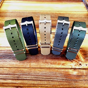 Pu&ograve; includere: Cinque cinturini per orologi in nylon di vari colori: verde, blu navy, beige, grigio e verde acqua. Ogni cinturino presenta una fibbia color argento e il testo "NATO-G10". I cinturini sono esposti su una superficie di legno.
