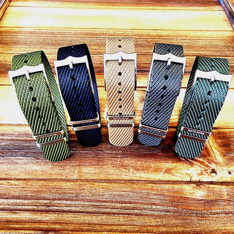 Pu&ograve; includere: Cinque cinturini per orologi in nylon di vari colori: verde, blu navy, beige, grigio e verde acqua. Ogni cinturino presenta una fibbia color argento e il testo "NATO-G10". I cinturini sono esposti su una superficie di legno.