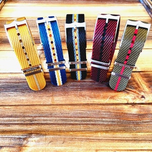 Pu&ograve; includere: Cinque cinturini per orologi in nylon di vari colori, tra cui giallo, blu, nero, rosso e verde oliva. Ogni cinturino presenta una fibbia argentata e il testo "NATO-G10e". I cinturini sono esposti su una superficie di legno.