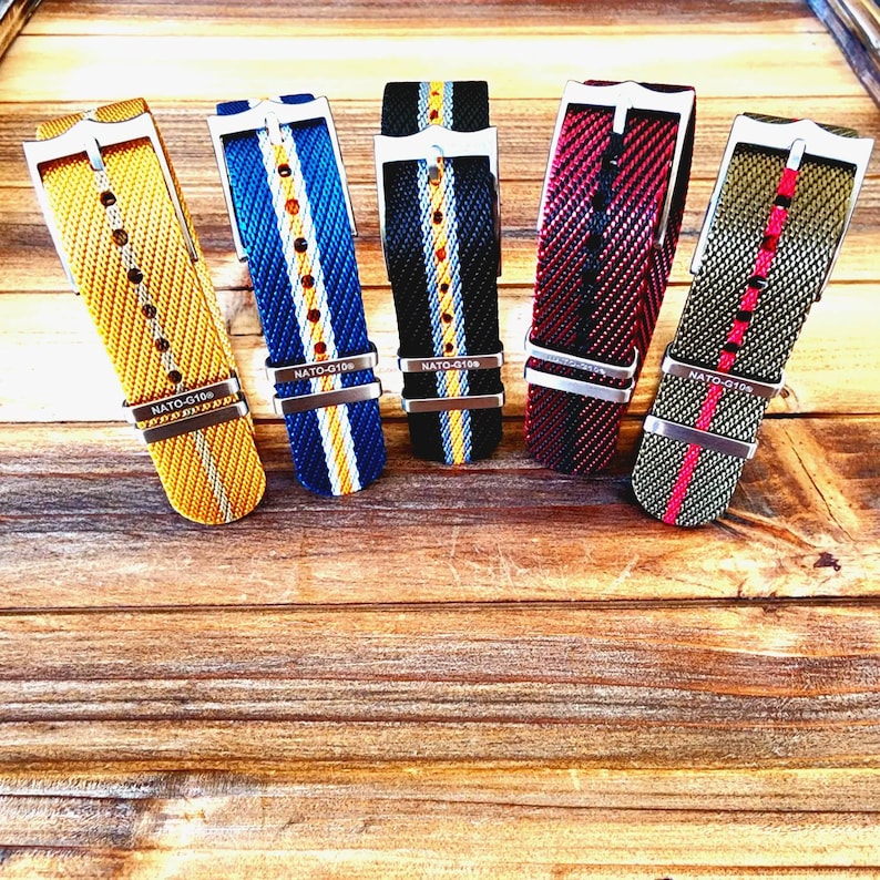 Pu&ograve; includere: Cinque cinturini per orologi in nylon di vari colori, tra cui giallo, blu, nero, rosso e verde oliva. Ogni cinturino presenta una fibbia argentata e il testo "NATO-G10e". I cinturini sono esposti su una superficie di legno.