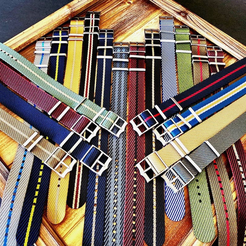 Pu&ograve; includere: Una collezione di cinturini colorati per orologi con vari disegni a righe e fantasia. I cinturini presentano una variet&agrave; di colori, tra cui blu, verde, rosso e giallo, con fibbie color argento. I cinturini sono disposti in un vassoio di legno.
