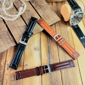 May include: Three leather watch straps in black, brown, and tan, with silver buckles. A wristwatch with a black leather band is visible in the background. The straps are arranged on a wooden surface.