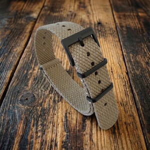 Puede incluir: Una correa de reloj de nailon beige con herrajes negros. La correa presenta una textura tejida y una hebilla negra. La marca "NATO-STRAP" es visible en la hebilla. La correa est&aacute; dise&ntilde;ada para un reloj.