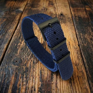 Puede incluir: Una correa de reloj azul marino con un tejido texturizado. La correa tiene una hebilla y presillas de metal negro. El texto "NATO-G10" es visible en la hebilla. La correa se muestra sobre una superficie de madera.