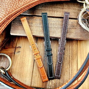 May include: Three leather watch straps in shades of brown, black, and tan, with white stitching. The straps are displayed on a wooden surface, with a leather bag and gold hardware in the background. The straps have silver buckles.