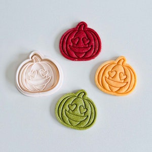 May include: Four pumpkin-shaped cookie cutters, each with a different color: red, orange, green, and white. The cutters have a smiling face with heart-shaped eyes.